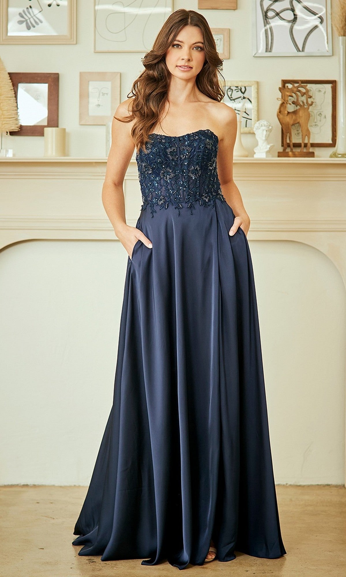 A woman stands indoors wearing the Amelia Couture 6204 long prom dress in navy blue, showcasing a strapless fitted lace beaded bodice, flowing A-line skirt, and convenient pockets.